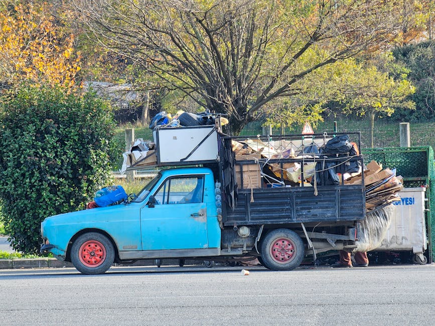 A small blue pickup truck parked on the side of a street, loaded with a variety of waste materials including large cardboard boxes, black plastic bags, and miscellaneous debris. The vehicle's body sho