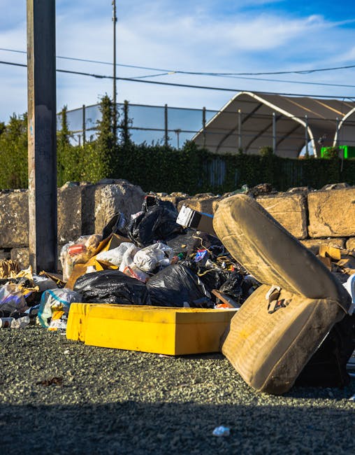 A small blue pickup truck parked on the side of a street, loaded with a variety of waste materials including large cardboard boxes, black plastic bags, and miscellaneous debris. The vehicle's body sho